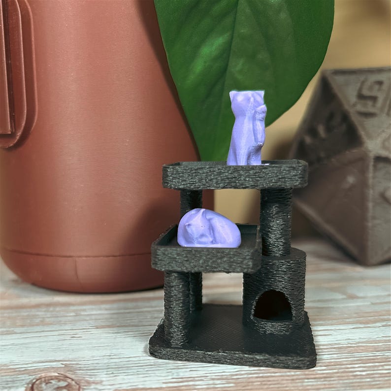 Miniature black cat tree with lavender sleeping and sitting cat figures, shown on wood surface with brown planter, green plant, and large dice decor in background