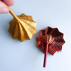 Two 3D printed star fidgets with layered, ridged designs shown on a light surface. One is metallic gold, and the other blends red and gold with a rainbow base. A hand holds the handle of the gold fidget in the upper left.