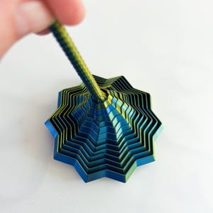 May include: A multi-layered, star-shaped object with a textured, cylindrical handle. The object features a gradient of blue and gold colors. The handle is held by a person's hand.
