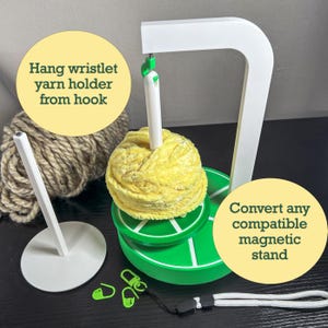 May include: A white yarn holder with a green base and a yellow yarn ball. The holder has a hook to hang a wristlet yarn holder. The text says, "Hang wristlet yarn holder from hook" and "Convert any compatible magnetic stand."