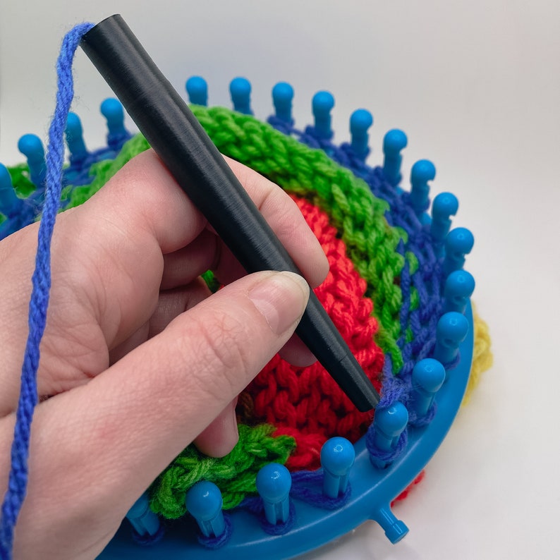 May include: A round blue plastic knitting loom with a series of upright pegs along the edge. A black knitting tool is positioned over the loom, which contains a partially completed circular knit project featuring vibrant red, green, and blue yarn.