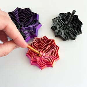 May include: Three colorful, layered star-shaped tops. One is purple and black, another is black, and the third is red and pink. A hand is inserting a gold-colored rod into the red and pink top. The tops are on a white surface.