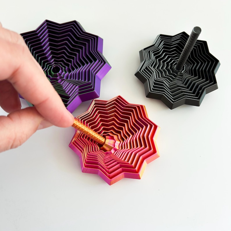May include: Three colorful, layered star-shaped tops. One is purple and black, another is black, and the third is red and pink. A hand is inserting a gold-colored rod into the red and pink top. The tops are on a white surface.