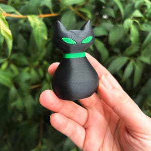May include: A black cat figurine with green eyes and a green collar. The cat has a stylized design and is held in a hand against a background of green leaves. The figurine is approximately 3 inches tall.
