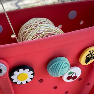 May include: A bright red tote bag with a ball of yarn inside. The bag has several decorative charms attached, including a daisy, a ball of yarn, a pair of cherries, and a bee. The yarn is a mix of yellow and pink.