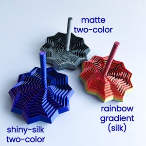 Three 3D printed star fidgets in different finishes: shiny silk blue/green, matte gray/red, and rainbow gradient silk. Each labeled with blue text describing its color style on a soft blue background.