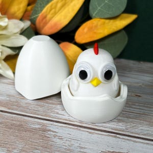 May include: A white ceramic chick hatching from a white ceramic egg. The chick has large black eyes and a red comb.