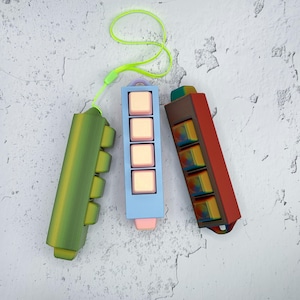 May include: Three rectangular macro keypads with four keys each. One is green with a neon yellow lanyard, one is light blue with pale pink keys, and one is a dark red and brown gradient with multi-colored keys featuring a swirling pattern.