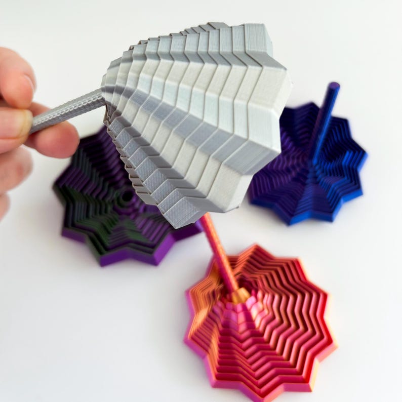 May include: A collection of colorful, geometric-shaped tops. One is held by a hand, showing a gray top with a zig-zag design. Other tops are in purple, blue, and red-orange gradients. The tops have a layered, star-like base.