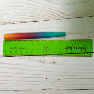 May include: A tapered cylindrical object with a smooth gradient finish transitioning from orange to red, purple, blue, and teal. It is positioned above a green plastic ruler, showing a total length of approximately 5.5 inches.