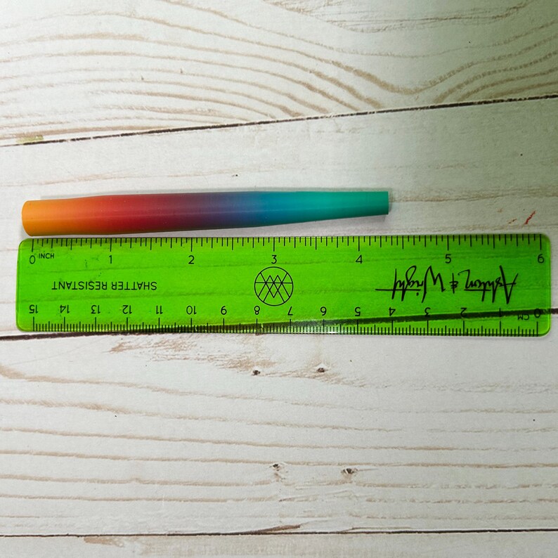 May include: A tapered cylindrical object with a smooth gradient finish transitioning from orange to red, purple, blue, and teal. It is positioned above a green plastic ruler, showing a total length of approximately 5.5 inches.