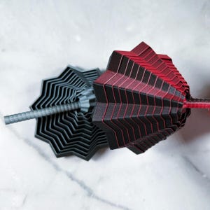 Close-up of two stacked 3D printed star fidgets in matte red/black and gray. The stepped, geometric layers create a hypnotic pattern against a light marble background.