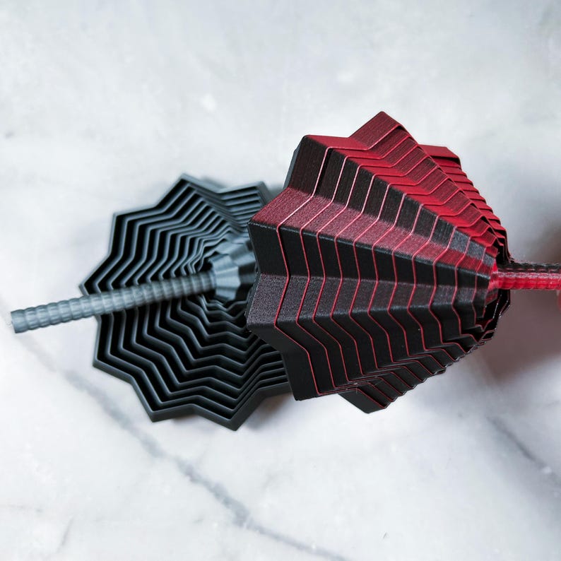 Close-up of two stacked 3D printed star fidgets in matte red/black and gray. The stepped, geometric layers create a hypnotic pattern against a light marble background.