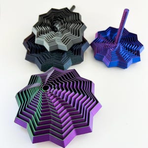 May include: A collection of 3D-printed geometric sculptures in various colors. The sculptures have a layered, star-like design with a central hole. Colors include black, gray, blue, purple, and green. One sculpture has a central rod.