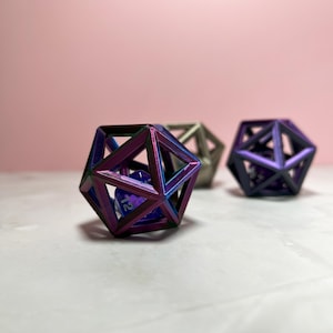 May include: Three iridescent metal dice cages, one with a purple and blue cage and a purple d12 die inside, one with a silver cage and a purple d12 die inside, and one with a purple cage and a blue d12 die inside.