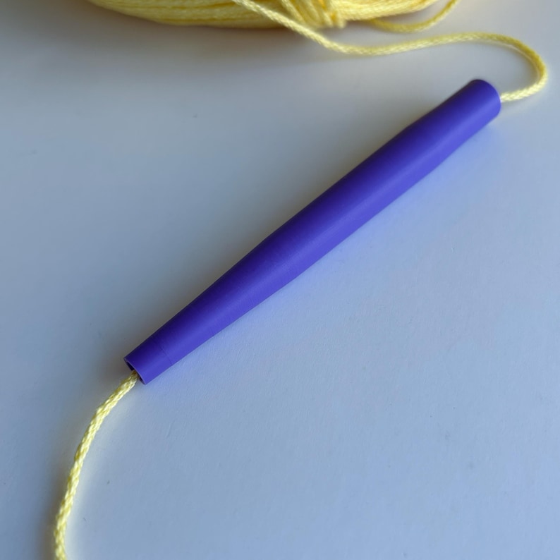 May include: A smooth, tapered purple crochet hook with a matte finish, threaded with a single strand of pale yellow yarn that extends from both ends of the tool, showcasing the contrast between the vibrant purple plastic and the light yellow fiber.