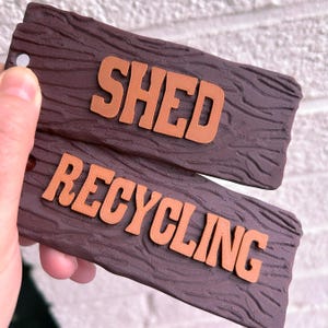 May include: Two wooden signs with a wood grain texture. The top sign says "SHED" in orange letters. The bottom sign says "RECYCLING" in orange letters.