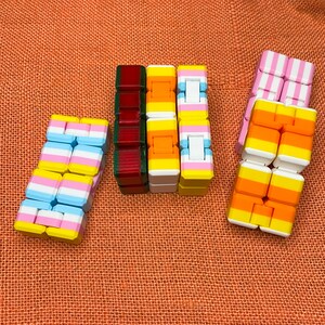 May include: A set of six colorful fidget toys, each with a different pattern of pink, blue, yellow, orange, and white squares. The toys are made of plastic and are designed to be squeezed and manipulated.