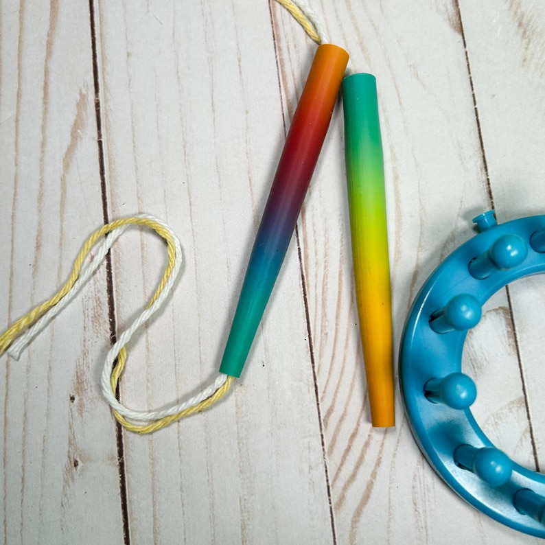 May include: Two tapered knitting needles with a gradient finish, one transitioning from orange to purple to teal and the other from teal to yellow to orange, alongside a blue circular knitting loom and a length of yellow and white twisted yarn.
