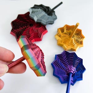 Hand holding a 3D printed rainbow star fidget with a ridged, layered surface. Other star fidgets in matte red/black, gold, purple/blue, and white and black rest on a white background, showing a range of available colors.