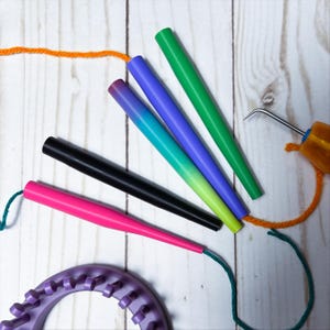 May include: A set of four smooth, tapered plastic knitting needles in bright green, purple, black, and pink, accompanied by a purple circular knitting loom and a metal hook tool with an amber handle, with orange and green yarn attached.
