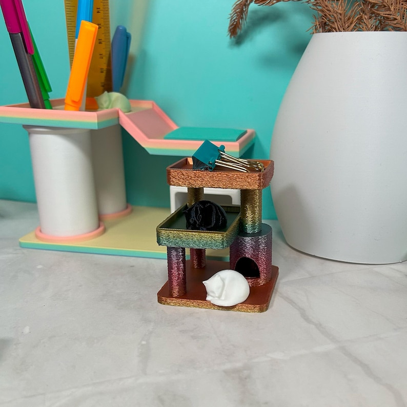 Miniature rainbow silk cat tree with small white and black cat figures; used as desk organizer with paper clips on platform; colorful desk accessories in background.