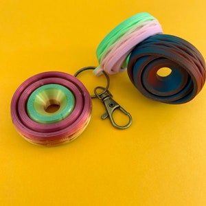 May include: A multi-colored, layered, circular keychain with a metal ring and a silver clasp. The keychain is made of a flexible material and has a green, pink, and brown color scheme.