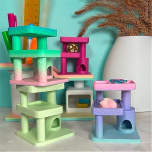 Colorful 3D printed cat tree desk buddies in mint green, bright pink, and pastel rainbow hues. These look like miniature cat furniture with platforms, scratching posts, and hideaway caves, displayed on white desk surface.