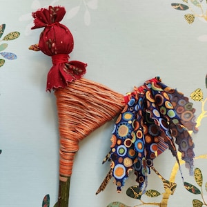 Puede incluir: Una figura de gallo hecha a mano, elaborada con un palo de madera, envuelta con hilo naranja y adornada con detalles de tela roja. La cola presenta una vibrante mezcla de telas estampadas en azul, naranja y amarillo. La cabeza del gallo es de tela roja.