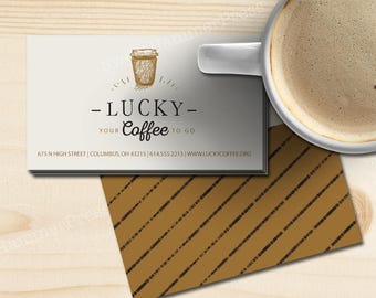 May include: A white business card with brown text and a drawing of a coffee cup. The text reads "Lucky Coffee" and includes the business's address, phone number, and website. The card is on a wood surface next to a white coffee mug.