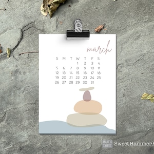 May include: A white March calendar page clipped to a metal clip. The calendar features a minimalist design with stacked stone art and the text "march" in a soft, handwritten font. The calendar is set against a textured gray background.