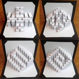 May include: Four white paper pop-up cards with different 3D geometric designs. Each card is framed with a black border and set against a wooden background.