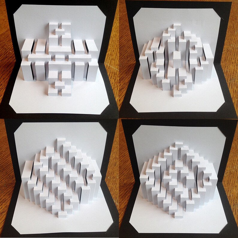 May include: Four white paper pop-up cards with different 3D geometric designs. Each card is framed with a black border and set against a wooden background.