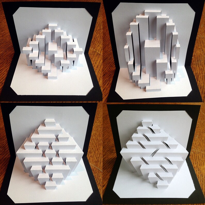 May include: Four white paper pop-up cards with different geometric designs. Each card is framed in black and set against a wood background.
