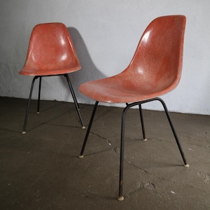 May include: Two mid-century modern chairs. The chairs have a molded, reddish-orange seat and a black metal base. The legs are thin and angled, with small white feet. The chairs are set against a white wall.
