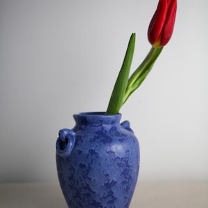 May include: A blue ceramic vase with a textured surface, featuring two small handles. A single red tulip with a green stem is placed inside the vase. The vase is on a neutral surface against a white background.