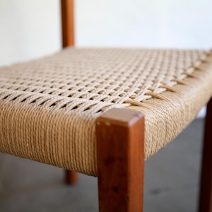 May include: A close-up of a wooden chair with a woven seat. The seat is made of a light brown rope material, and the frame is a darker brown wood. The chair has a simple, minimalist design, with clean lines and a natural aesthetic.