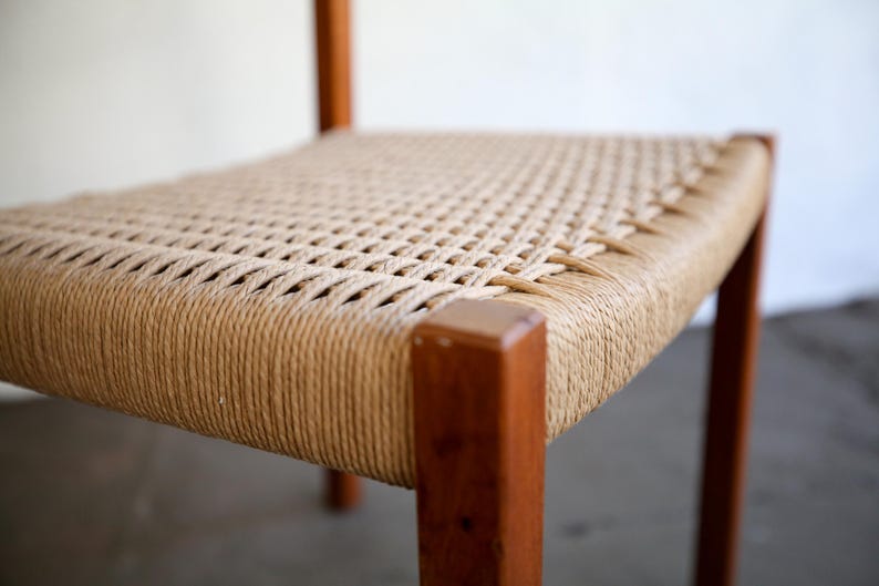 May include: A close-up of a wooden chair with a woven seat. The seat is made of a light brown rope material, and the frame is a darker brown wood. The chair has a simple, minimalist design, with clean lines and a natural aesthetic.