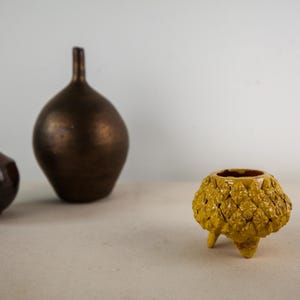 May include: A small yellow tripod vessel with a textured, repeating floral pattern on its exterior, accompanied by a dark brown spherical vase and a smaller, rounded vase featuring a glossy finish in shades of deep red and dark brown.