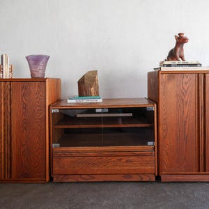 May include: A set of three wooden storage cabinets with a rich brown wood grain finish. The two taller cabinets feature solid doors, while the central unit includes a glass-fronted display section with internal shelving and a lower drawer.