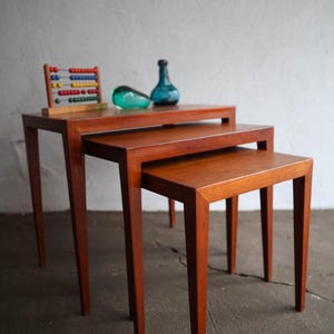 May include: Set of three nesting tables with a warm wood finish. The tables have a simple, rectangular design with tapered legs. Decorative glass bottles and a colorful abacus sit on top of the tables.