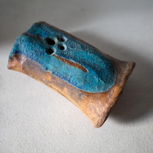 May include: A rectangular ceramic object with a textured, light brown base and a vibrant, raised blue glaze on the top surface. The blue section features a distinct, elongated shape with four small, circular holes arranged in a cluster near one end.