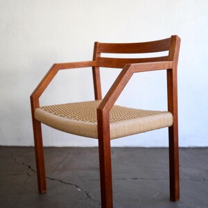 May include: A mid-century modern armchair with a woven seat and armrests. The chair features a solid wood frame with a natural finish. The seat is made of woven natural fibers. The chair is designed with a minimalist aesthetic.
