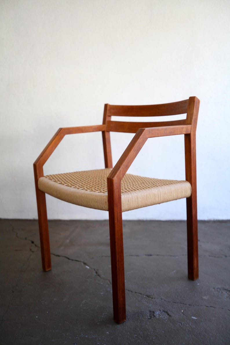 May include: A mid-century modern armchair with a woven seat and armrests. The chair features a solid wood frame with a natural finish. The seat is made of woven natural fibers. The chair is designed with a minimalist aesthetic.