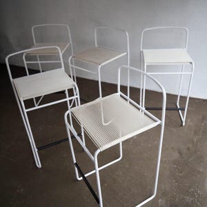 May include: A collection of five white metal bar stools. The seats vary, with some featuring light wood and others woven cream-colored material. The stools have a minimalist design with black support bars, suitable for a modern kitchen or bar setting.