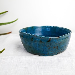 May include: A teal blue ceramic bowl with a rustic, uneven glaze. The bowl has a slightly rounded, rectangular shape and is perfect for serving snacks or small dishes.