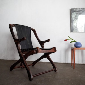 May include: A dark wood chair with a black leather seat and back. The chair has a unique, curved design. A small, square, wooden side table with a blue vase and a red flower sits nearby. A framed print hangs on the white wall.