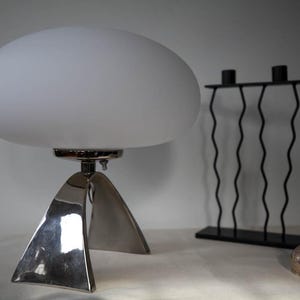 May include: A modern table lamp with a white, oval-shaped shade and a polished silver base. A black metal candle holder and a silver vase are also in the image. The lamp's base has a unique, geometric design. The objects are on a white surface.