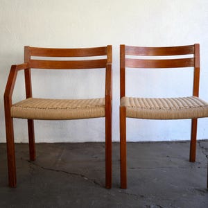 May include: Two wooden chairs with woven seats. One chair has armrests. The chairs have a light brown woven seat and a dark brown wooden frame. The chairs are set against a white wall and a dark gray floor.