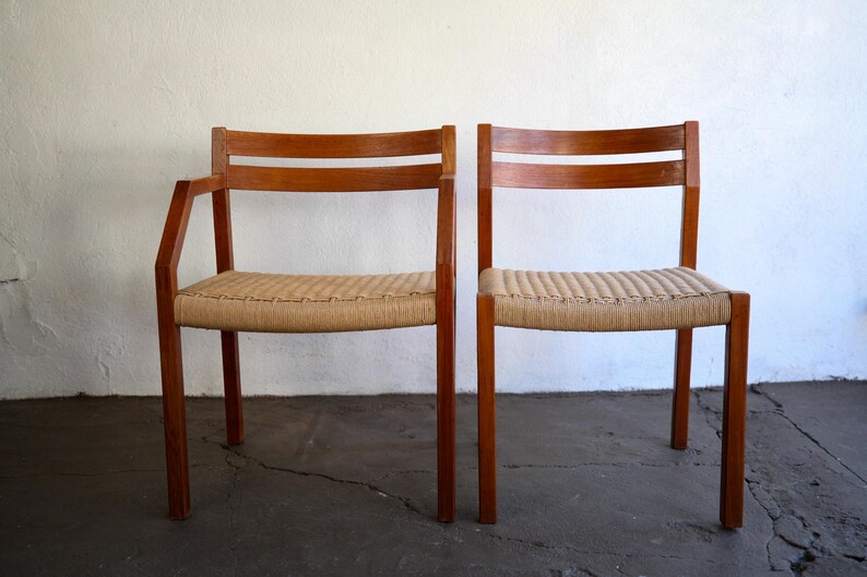 May include: Two wooden chairs with woven seats. One chair has armrests. The chairs have a light brown woven seat and a dark brown wooden frame. The chairs are set against a white wall and a dark gray floor.